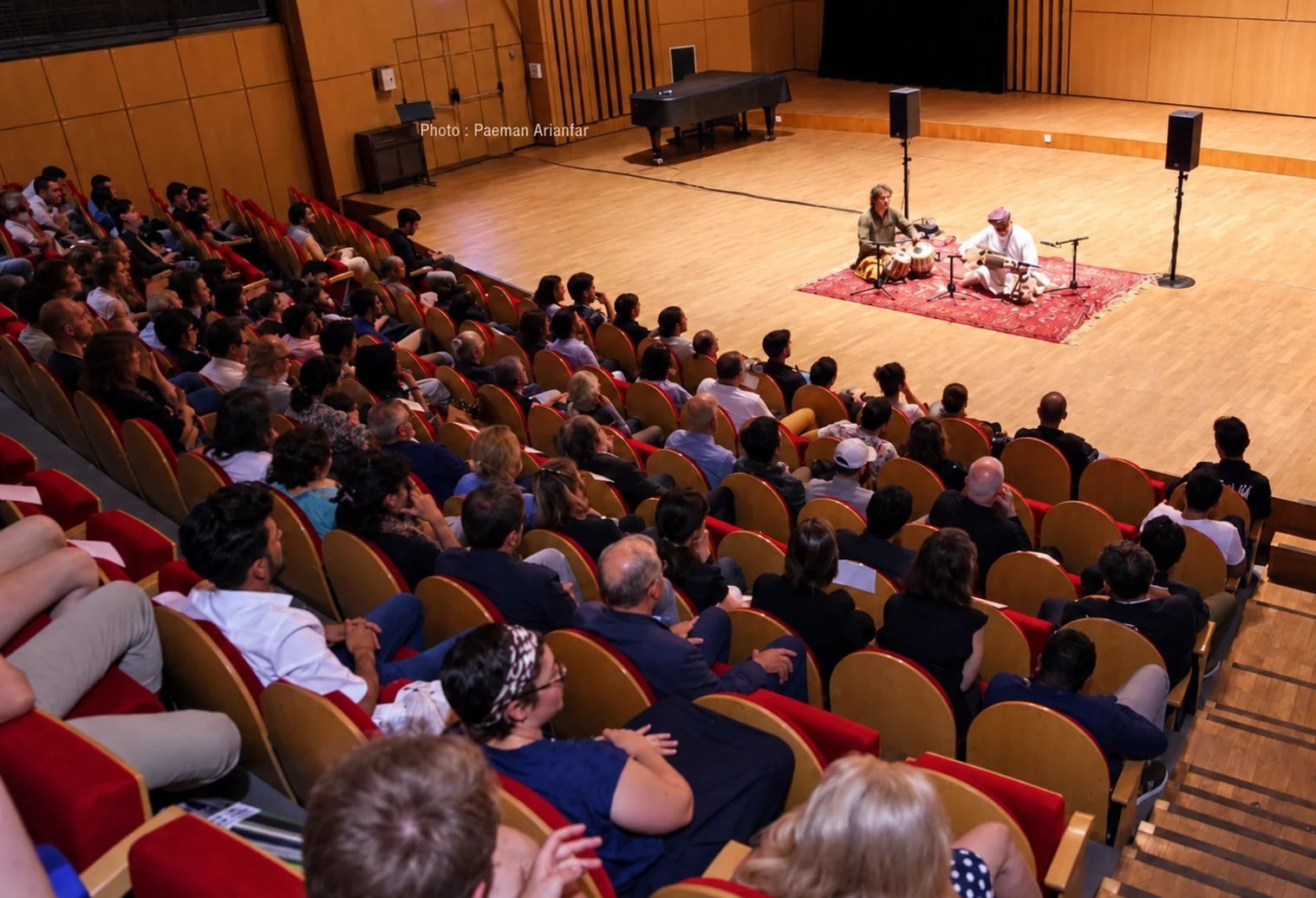 Concert de musique classique afghane : deux musiciens assis sur un tapis, rubab et tabla, devant une salle comble